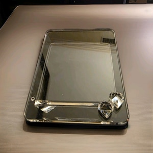 May include: A rectangular mirrored tray with a smooth, reflective surface and beveled edges. The tray features a clear glass accent bar across the lower section, adorned with two faceted, teardrop-shaped crystal ornaments on the right side.