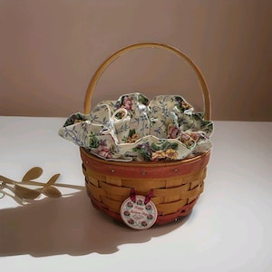 May include: A round woven wooden basket with a single arched handle and a fabric liner featuring a floral pattern in shades of pink, yellow, and green. A circular tag hangs from the front with the text Happy Mothers Day 1998 in red script.