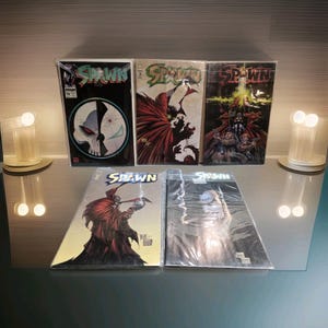 Op de afbeelding: Een verzameling van vijf Spawn-stripboeken in beschermende plastic hoezen. Drie staan rechtop met diverse karakterillustraties en het Spawn-logo. Twee liggen plat aan de voorzijde en tonen gedetailleerde kunstwerken van de figuur met cape.