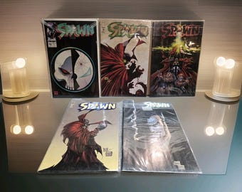 May include: A collection of five Spawn comic books in protective plastic sleeves. Three are standing upright, featuring various character illustrations and the Spawn logo. Two are lying flat in front, displaying detailed artwork of the caped figure.