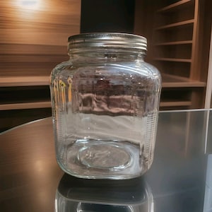 Puede incluir: Un frasco de almacenamiento de vidrio transparente de forma cuadrada con una boca ancha y una tapa metálica de rosca. El vidrio presenta una textura sutil con nervaduras verticales en las esquinas, lo que aporta estructura al recipiente.