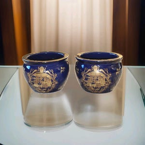 Peut inclure: Une paire de petits pots en céramique bleu cobalt, de forme arrondie, ornés d'un motif décoratif doré. Chaque pot présente une ouverture large avec un bord doré et une illustration dorée détaillée représentant des personnages.