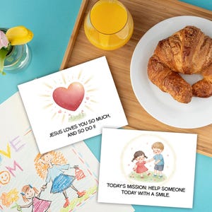 Pode incluir: Uma cena de café da manhã com um croissant em um prato branco, um copo de suco de laranja e cartões de felicitações. Um cartão diz: "Jesus ama você tanto, e eu também!" Outro diz: "A missão de hoje: ajude alguém hoje com um sorriso."
