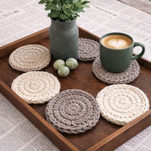 May include: A wooden tray holds a green mug of coffee with latte art, a green vase with greenery, and several crocheted coasters in shades of beige and gray. Decorative speckled eggs are also on the tray.