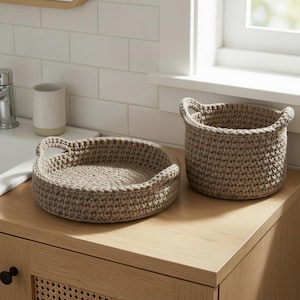 May include: Two beige crochet baskets with handles sit on a wooden cabinet. One is a shallow, round tray, and the other is a taller, cylindrical basket. The baskets are made of a thick, textured yarn and are placed near a window and a sink.