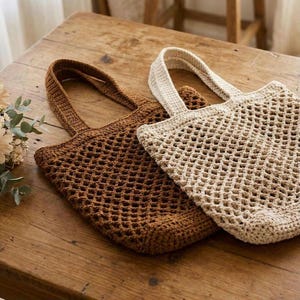 Può includere: Due borse tote all'uncinetto. Una è marrone caldo, l'altra beige chiaro. Entrambe le borse hanno un design a rete e manici robusti. Sono appoggiate su una superficie di legno.