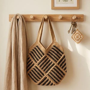 May include: A beige and black crocheted tote bag with a geometric pattern hangs on a wooden wall rack. The bag has two handles and a square shape. A beige scarf hangs on the left side of the rack. A set of keys and a crocheted square are also hanging on the rack.