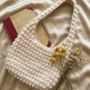 May include: Cream-colored crochet shoulder bag with a textured, bobble stitch design. The bag has a rounded shape and a single strap. A vintage book with the words "HANDMADE TREASURES" is visible. Yellow billy buttons and white daisies are arranged near the bag.
