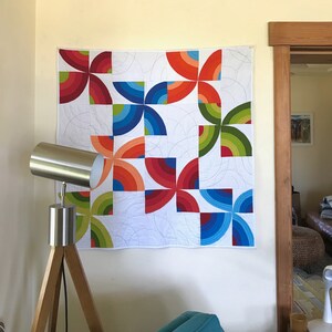 May include: A colorful quilt with a geometric pattern of overlapping circles in shades of red, orange, yellow, green, and blue. The quilt is hanging on a white wall.