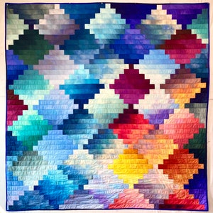 Pode incluir: Um cobertor colorido com um padrão geométrico de quadrados e retângulos em tons de azul, roxo, vermelho, laranja e amarelo. O cobertor é feito de tecido que foi tingido em um padrão degradê, criando um design único e atraente.