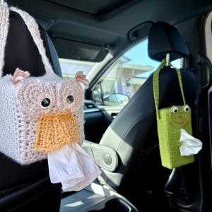 May include: Crocheted tissue box covers in the shape of an owl and a frog. The owl cover is light gray with large googly eyes and an orange beak. The frog cover is green with a smiling face. Both are hanging in a car.
