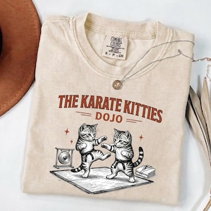 May include: A beige short-sleeved t-shirt featuring a graphic of two kittens wearing black belts and practising karate on a mat. The text reads THE KARATE KITTIES DOJO. A small gong and a folded white garment are visible on the mat near the kittens.