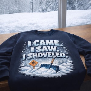 Peut inclure: Sweat-shirt bleu marine avec l'inscription "I CAME. I SAW. I SHOVELED." Le motif représente une pelle, un panneau routier et un paysage enneigé. Le sweat-shirt est posé sur une surface en bois, avec une vue enneigée en arrière-plan.