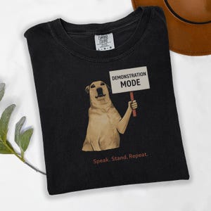Może przedstawiać: Czarny t-shirt z grafiką psa trzymającego znak z napisem "DEMONSTRATION MODE". Poniżej psa znajduje się napis "Speak. Stand. Repeat." Koszulka marki Comfort Colors.