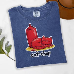 Puede incluir: Camiseta azul con una ilustración de dibujos animados de un gato rojo y una botella de ketchup. El gato está tumbado en un círculo amarillo, con el texto "cat-chup" debajo.