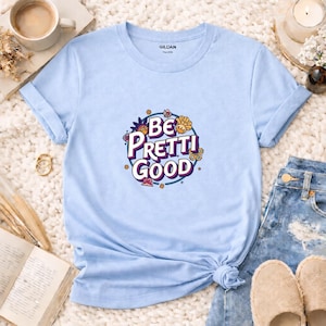 May include: Pale blue t-shirt with rolled sleeves and a knotted hem. The shirt displays the text "Be Pretti Good" in a circular design with floral and butterfly accents. The Gildan brand logo is visible on the shirt.