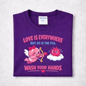 Può includere: Una maglietta viola con la scritta "LOVE IS EVERYWHERE BUT SO IS THE FLU, WASH YOUR HANDS". La maglietta presenta un cupido dei cartoni animati con arco e freccia e un virus dei cartoni animati su una nuvola.