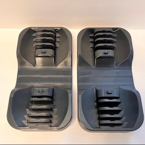 May include: Two dark gray, 3D-printed objects. Each has a curved, rectangular base with a series of stacked, rectangular platforms and a central, ridged section. The objects are designed with a series of grooves and cutouts.