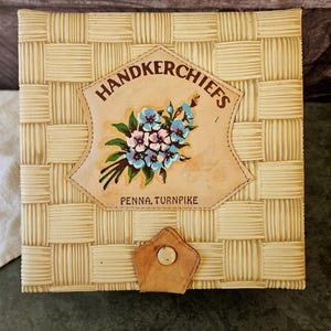 May include: A square, woven-patterned box in a light beige color. The box features a leather patch with the word "HANDKERCHIEFS" and a floral design. The text "PENNA. TURNPIKE" is below the floral design. A leather clasp with a button secures the box.