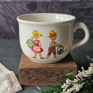 May include: Cream-colored ceramic mug with a whimsical illustration of a girl holding flowers and a boy with a watering can. The mug has a rounded shape and a large handle. The design features bright colors and a vintage aesthetic.