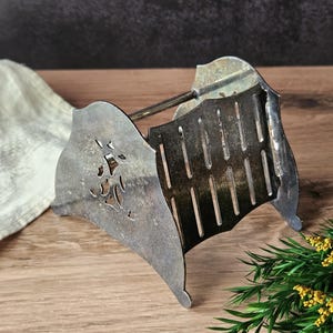May include: An antique silver-toned toast rack with a decorative floral design on the side panels. The rack has a series of vertical slots for holding toast and a handle at the top. The rack is placed on a wooden surface.