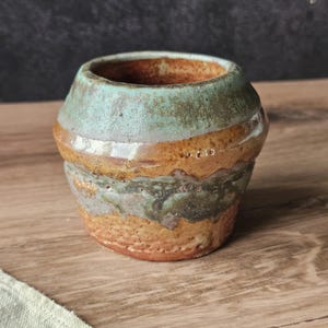 May include: A small, round ceramic cup featuring a textured, multi-colored glaze. The upper portion is a muted sage green, transitioning into a warm, earthy ochre and brown band in the middle, with a speckled, rough-textured base in tan.