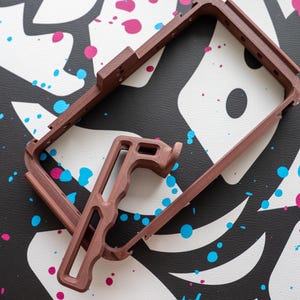 May include: A brown, rectangular camera cage with a handle, designed to securely hold a camera. The cage has multiple mounting points and is made of a durable material. The background is a black and white pattern with blue and pink paint splatters.
