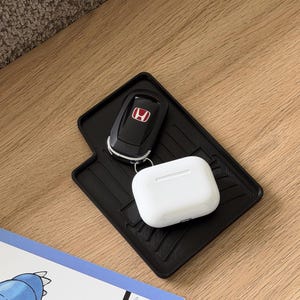 May include: A black car key with a red Honda logo and a white wireless earbud case rest on a black rubber mat. The items are on a light brown wooden surface.