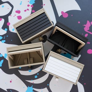 May include: Four miniature garage door models in beige, black, and white, with varying door designs. The models are arranged on a surface with a black, white, and pink abstract design.