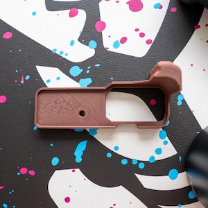 May include: A brown, rectangular 3D-printed object with a handle-like extension. The object has a small circular hole and a textured surface. It rests on a black surface with pink and blue paint splatters.