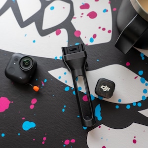 May include: A black action camera, a black handle, and a small black square device with the DJI logo are on a black surface with white and colourful paint splatters. A black mug with coffee is in the background.