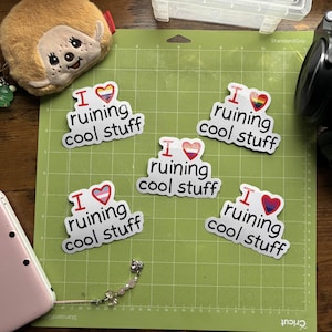 May include: Five white stickers with the text "I ruining cool stuff" in a black, bold font. Each sticker has a different heart design in the upper right corner. The stickers are on a green cutting mat.