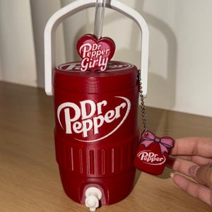 K&ouml;nnte beinhalten: Ein roter, zylindrischer Getr&auml;nkek&uuml;hler mit wei&szlig;em Griff und Zapfhahn, der das Dr Pepper-Logo tr&auml;gt. Ein durchsichtiger Strohhalm ragt oben heraus, verziert mit einem herzf&ouml;rmigen Anh&auml;nger mit der Aufschrift Dr Pepper Girly. Ein kleiner Dr Pepper-Anh&auml;nger mit rosa Schleife h&auml;ngt an einer Kette.