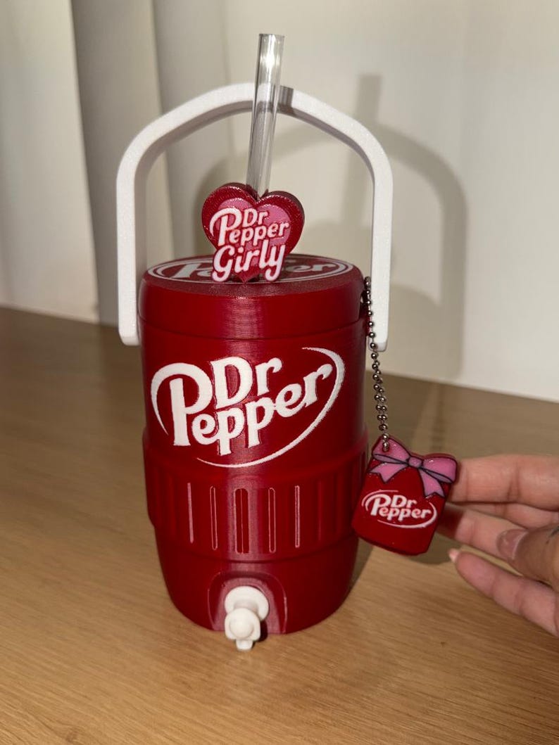K&ouml;nnte beinhalten: Ein roter, zylindrischer Getr&auml;nkek&uuml;hler mit wei&szlig;em Griff und Zapfhahn, der das Dr Pepper-Logo tr&auml;gt. Ein durchsichtiger Strohhalm ragt oben heraus, verziert mit einem herzf&ouml;rmigen Anh&auml;nger mit der Aufschrift Dr Pepper Girly. Ein kleiner Dr Pepper-Anh&auml;nger mit rosa Schleife h&auml;ngt an einer Kette.