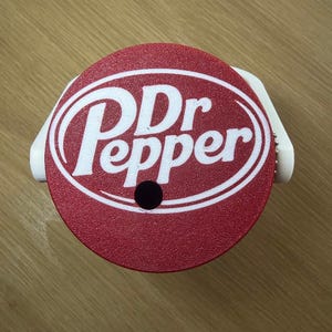 K&ouml;nnte beinhalten: Ein runder roter Kunststoffdeckel mit dem wei&szlig;en Dr Pepper-Logo in der Mitte. Der Deckel hat ein kleines kreisf&ouml;rmiges Loch nahe dem unteren Rand und eine wei&szlig;e Kunststoffhalterung an den Seiten, die f&uuml;r einen Getr&auml;nkebeh&auml;lter gedacht ist.