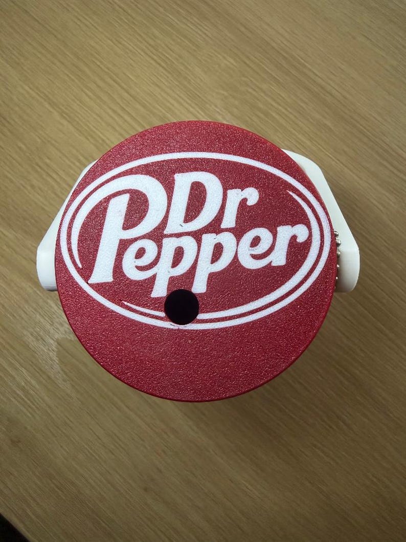 K&ouml;nnte beinhalten: Ein runder roter Kunststoffdeckel mit dem wei&szlig;en Dr Pepper-Logo in der Mitte. Der Deckel hat ein kleines kreisf&ouml;rmiges Loch nahe dem unteren Rand und eine wei&szlig;e Kunststoffhalterung an den Seiten, die f&uuml;r einen Getr&auml;nkebeh&auml;lter gedacht ist.