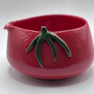 May include: A glossy red ceramic bowl shaped like a tomato, featuring a small pouring spout on the left side and a raised, dark green stem detail attached to the front. The surface has a smooth, speckled texture with a rounded, bulbous form.