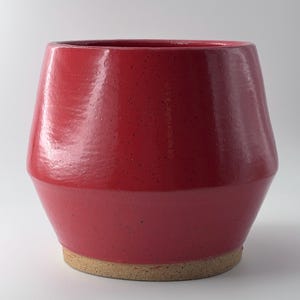 May include: A red ceramic planter with a textured surface and a natural, unglazed base. The planter has a wide opening and a slightly tapered shape, suitable for indoor or outdoor use. The red color is vibrant and eye-catching.