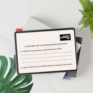 May include: A digital tablet displaying a workbook page with the text "CLARITY WORKBOOK CO." and prompts to answer questions about goals and time commitment. The tablet is on a stack of books and a green leaf.