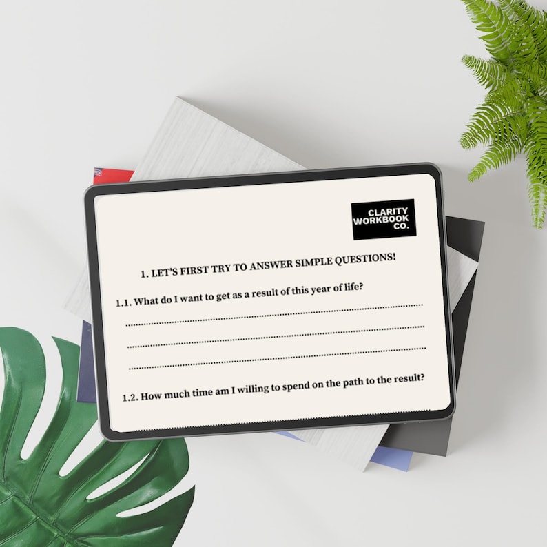 May include: A digital tablet displaying a workbook page with the text "CLARITY WORKBOOK CO." and prompts to answer questions about goals and time commitment. The tablet is on a stack of books and a green leaf.
