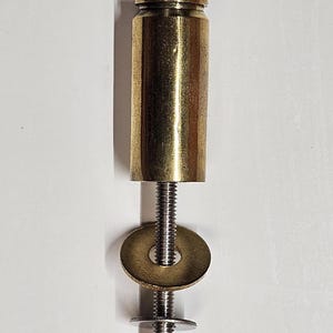 May include: A brass-colored furniture leg with a cylindrical top, threaded rod, and a washer. The leg is attached to a silver-colored bolt. The hardware is designed for furniture or other DIY projects.