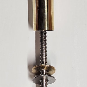 May include: A brass-colored, cylindrical object with a threaded rod, washers, and a nut. The top portion is a solid brass cylinder. The threaded rod has a gold-colored washer, a silver-colored washer, and a nut at the bottom.
