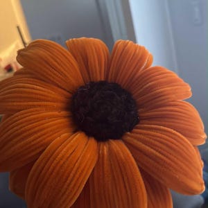 Puede incluir: Una gran flor de girasol de terciopelo naranja con un centro marrón oscuro. La flor tiene una textura detallada de pétalos y es una pieza decorativa vibrante. La imagen se centra en el diseño y el color de la flor.