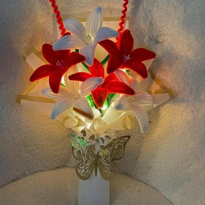 Puede incluir: Un arreglo floral decorativo con flores rojas y blancas con forma de lirio, iluminado con luz suave. Una mariposa dorada acentúa la base. La palabra "Wednesday" está escrita en cursiva en la parte superior.