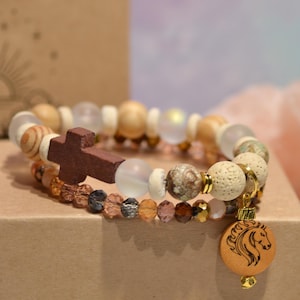 Op de afbeelding: Twee armbanden met houten kralen, een houten kruis en een paardenhanger. De armbanden hebben een verscheidenheid aan kralen in neutrale tinten, waaronder houten, heldere en patroon kralen. De armbanden worden op een bruine doos getoond.