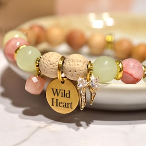 Puede incluir: Una pulsera de cuentas con una mezcla de cuentas verde claro, rosa y de madera. La pulsera presenta un dije dorado con las palabras "Wild Heart" y un pequeño dije de lazo. Una pequeña botella está en el fondo.
