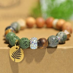 May include: A beaded bracelet featuring a variety of natural stone beads in shades of green, brown, and tan. A gold-colored charm with a fern design hangs from the bracelet. The bracelet includes a central bead with sparkling crystals.