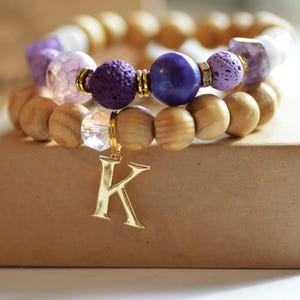 Op de afbeelding: Twee armbanden met houten en edelstenen kralen. Eén armband heeft een gouden bedel met de letter "K". De kralen zijn in tinten paars, blauw en wit. De armbanden worden gepresenteerd op een bruine doos.