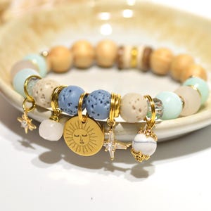 May include: A bracelet with wooden, blue, and beige beads, and gold charms. The charms include a sun, stars, and a globe. An amber essential oil bottle is next to the bracelet on a white surface.