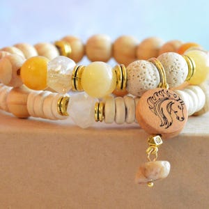 Op de afbeelding: Twee gestapelde armbanden met houten kralen, gele en heldere edelstenen en gouden accenten. Eén armband heeft een houten bedel met een paardenkopontwerp en een bungelende steen. De armbanden worden op een bruin oppervlak weergegeven.